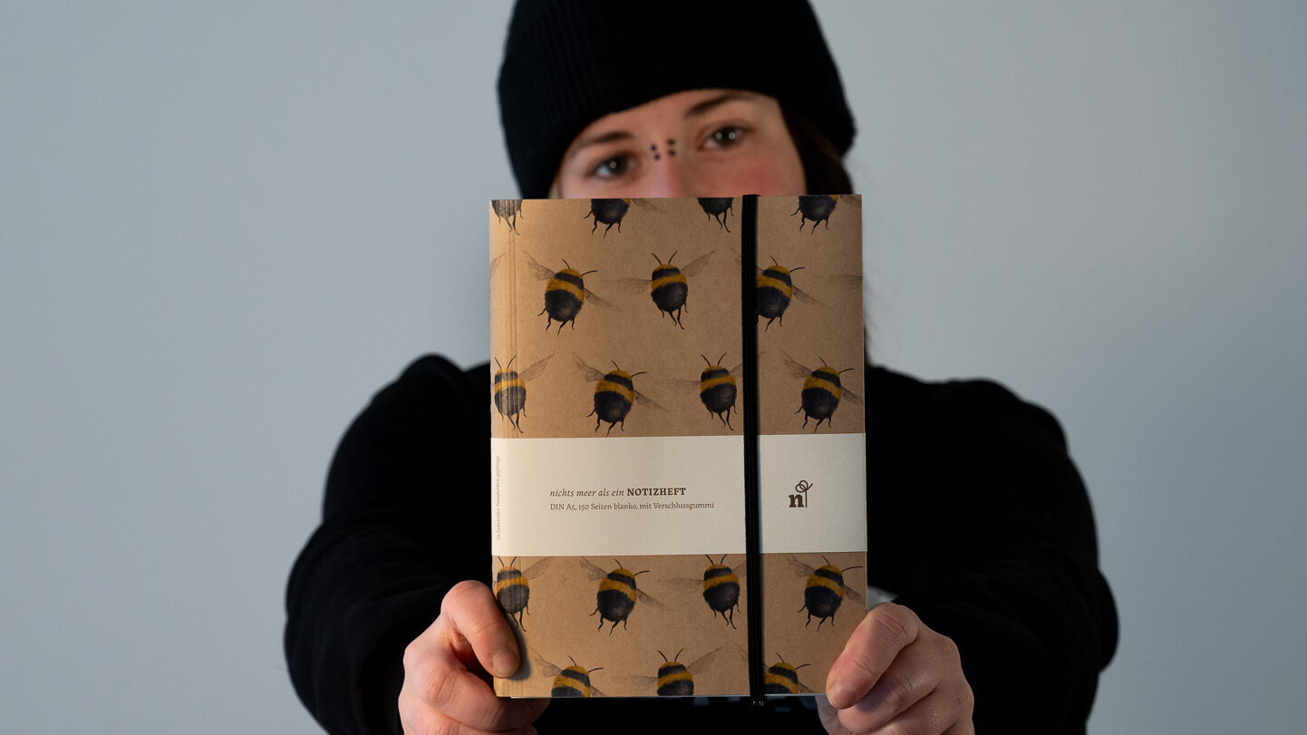 Ein Frau mit Pircings und Mütze hält ein Notizbuch mit Bienen vor ihr Gesicht.