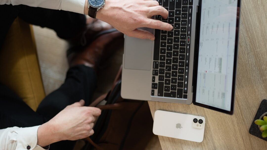 Ein Symbolbild: Ein Laptop steht auf dem tisch, ein iPhone liegt danaben. Die Hand einer vor dem Laptop sitzenden Person berührt die Tastatur. Das Bild ist eine Top Down Ansicht.