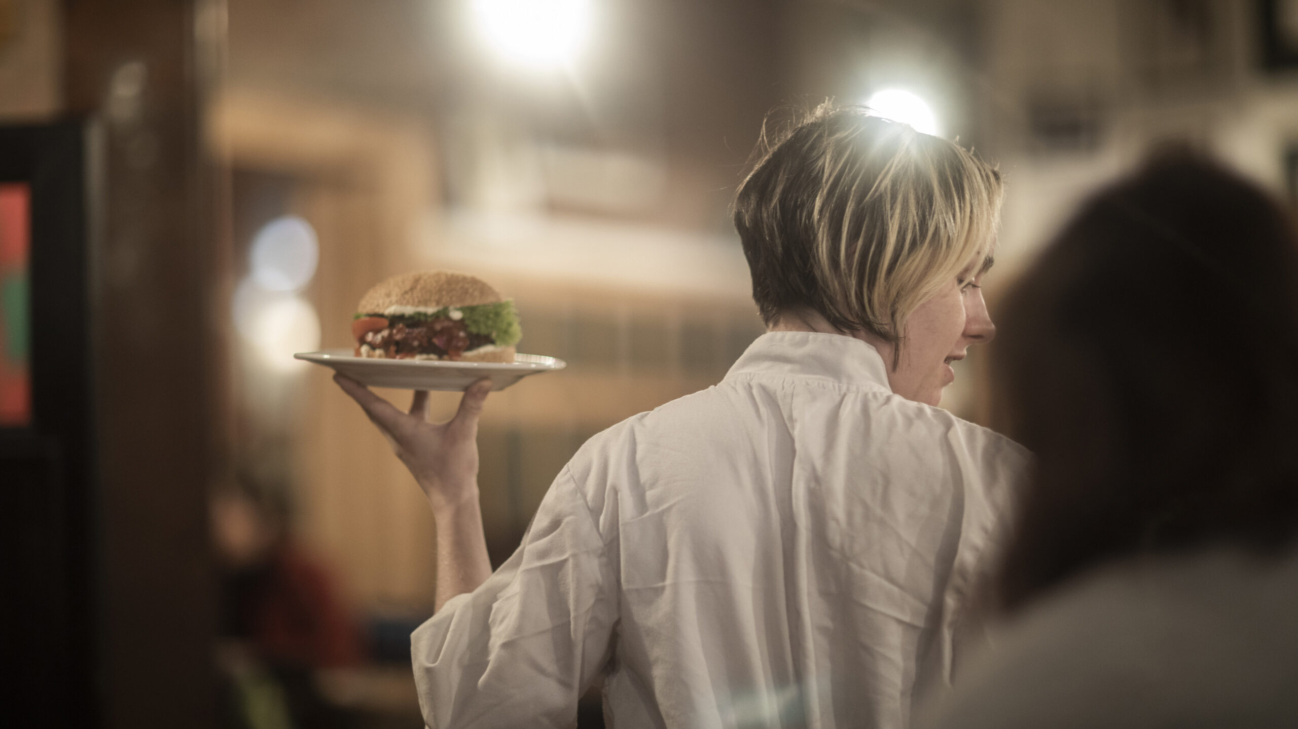Ein Symbolbild: Eine Frau (Servicekraft) trägt einen Burger. Man sieht sie von hinten und sie schaut zur Seite.